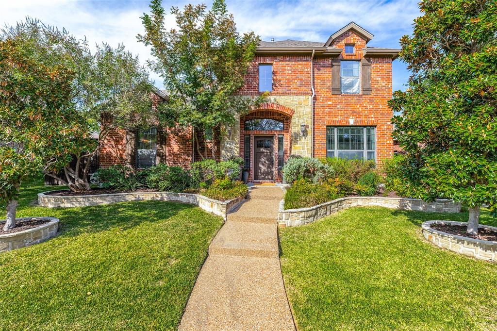Two-story brick and stone facade featuring an arched entryway, shingle roof, multi-pane windows, and stone-bordered landscaping beds