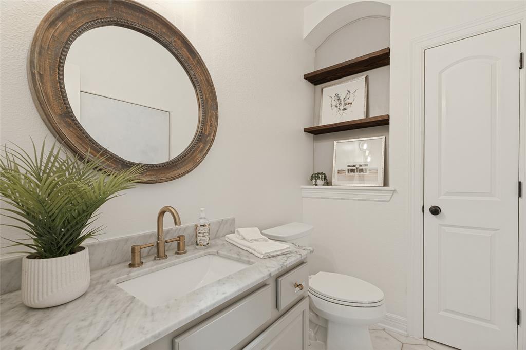 Vanity with stone countertop, undermount sink, and brass-finish widespread faucet, complemented by a circular wood-framed wall mirror, arched wall niche with wood shelving, and a paneled interior door linen closet
