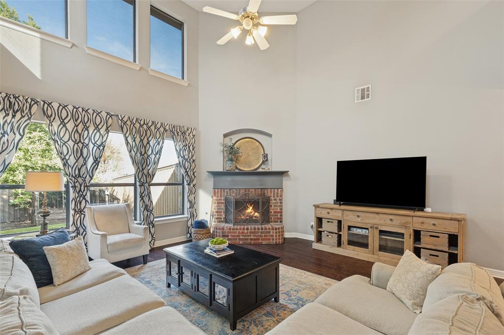 High-ceilinged living area featuring wood-finish flooring, floor-to-ceiling windows, a corner brick fireplace, and a ceiling fan