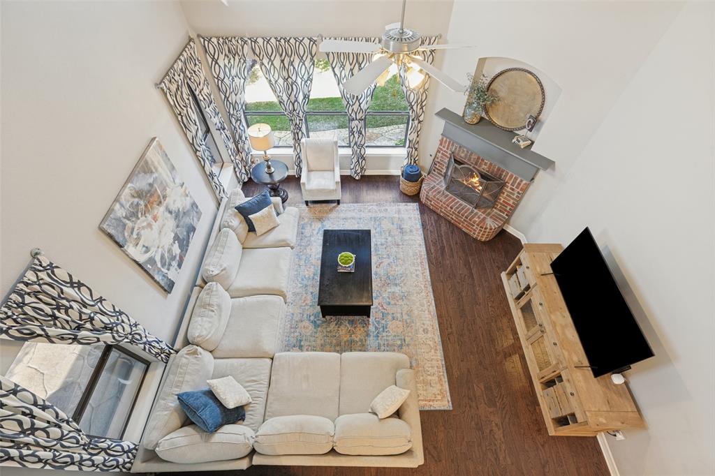 High-ceiling living area featuring wood-finish flooring, a corner brick fireplace with a mantel, and oversized windows