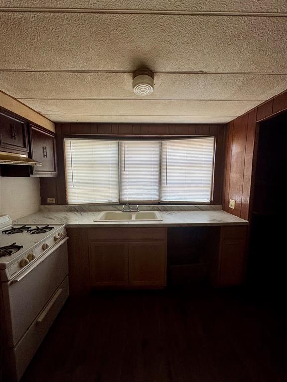 Kitchen featuring wood-finish cabinetry, stone-patterned laminate countertops, dual-basin sink with a chrome faucet, integrated gas range, and a three-pane window with horizontal blinds