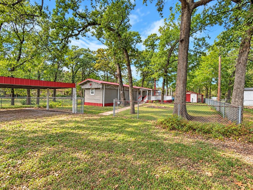 Detached carport with metal roofing, chain-link fencing, mature trees, and storage structure on a grass-covered lot