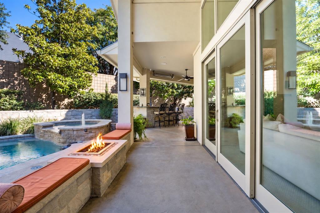 Covered patio featuring floor-to-ceiling glass panel doors, a built-in stone-veneer fire feature, an integrated swimming pool with fountain, and a recessed outdoor kitchen bar
