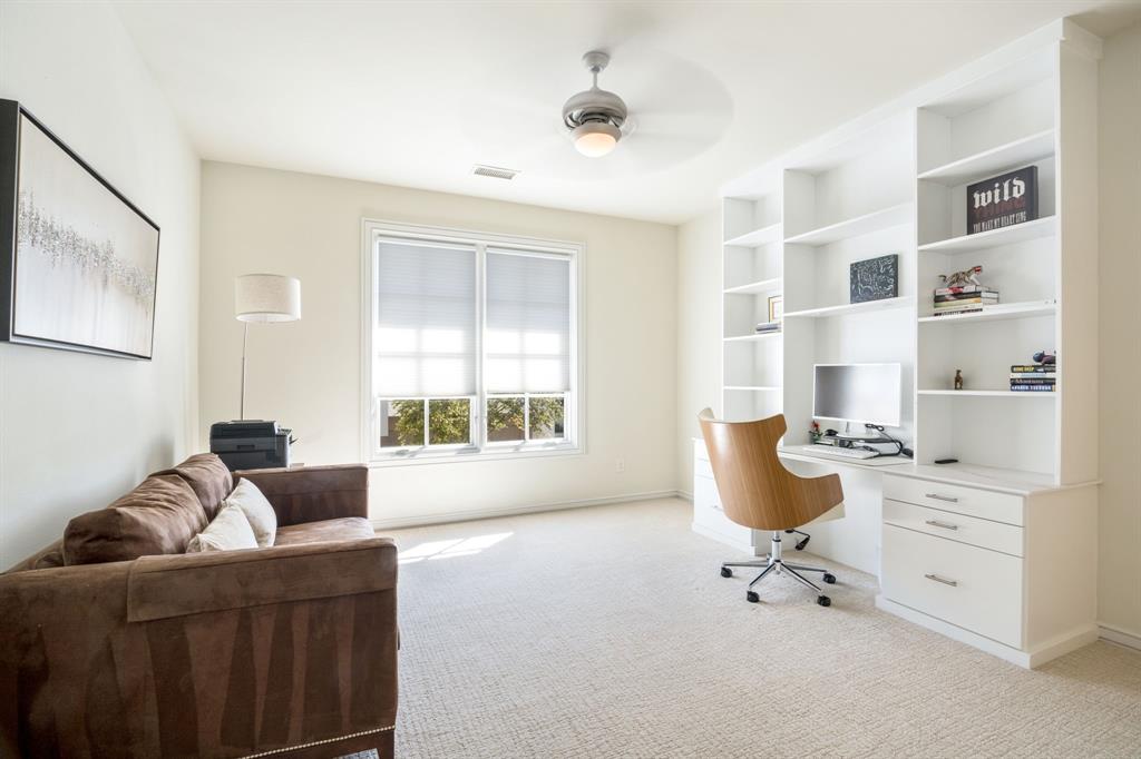 Built-in floor-to-ceiling shelving with integrated desk and drawer storage, neutral carpeting, dual-pane window with roller shade, and ceiling fan with lighting