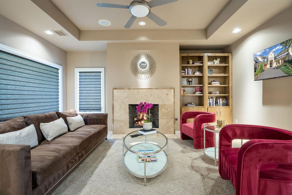Tray ceiling with recessed lighting, stone-surround fireplace, built-in wooden shelving, and dual windows with fitted shades