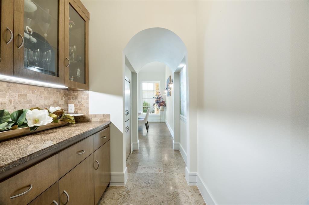 Built-in cabinetry with glass-front display uppers, stone-finish countertops, tiled backsplash, arched entryway, and natural stone flooring