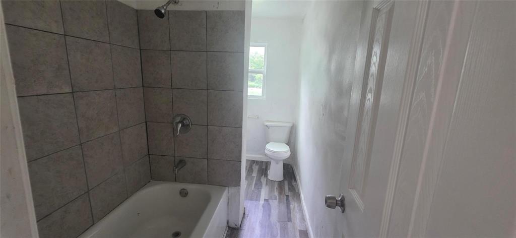 Bathroom featuring a porcelain tub with a chrome shower fixture, tiled surround, wood-finish flooring, and a standard toilet