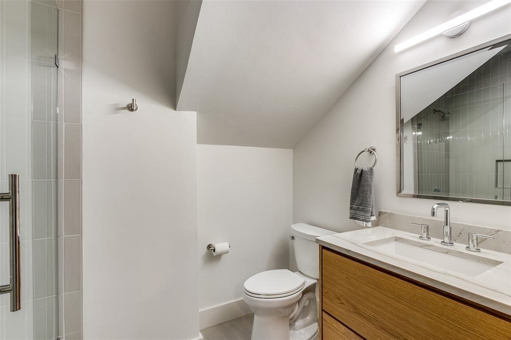 Bathroom featuring a wood-finish vanity with a stone-surface countertop, rectangular undermount sink, wall-mounted mirror with integrated lighting, and a glass-enclosed shower