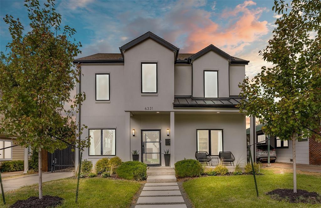 Two-story exterior featuring a neutral stucco facade, a metal roof accent, large rectangular windows with dark trim, a covered entryway, and a paved walkway