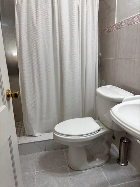 Bathroom featuring light-toned tile flooring, white porcelain fixtures, and a walk-in shower area with decorative accent tiling