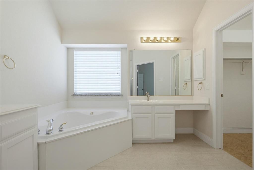 Corner soaking tub featuring a deck-mounted chrome faucet, white cabinetry with a built-in vanity, large vanity mirror, and wall-mounted light fixture