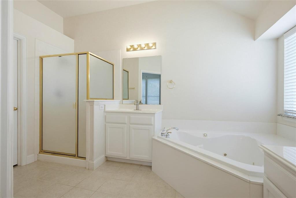 Bathroom featuring a jetted soaking tub, a separate walk-in shower with a gold-tone frame, a white vanity cabinet, a wall-mounted light fixture, and tile flooring