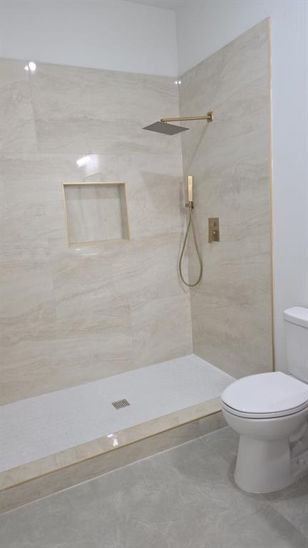 Walk-in shower featuring large-format stone-finish tile surround, a built-in recessed niche, gold-toned rainfall showerhead with handheld sprayer, and a low-profile basin