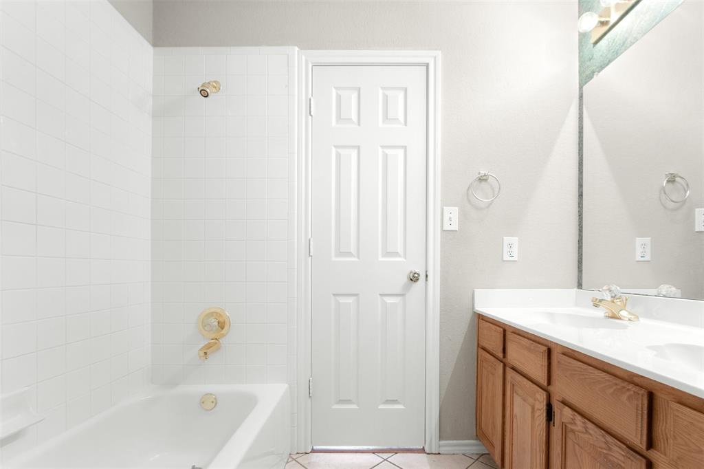 Bathroom featuring a dual-basin vanity with wood-finish cabinetry, white solid-surface countertop, gold-toned hardware, and a tile-surround shower and tub combination