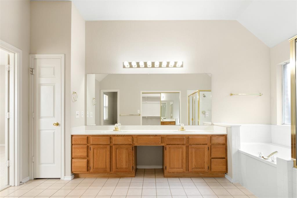 Dual-vanity cabinetry with a central seated vanity area, expansive wall-to-wall mirror, multi-bulb overhead lighting, and built-in soaking tub