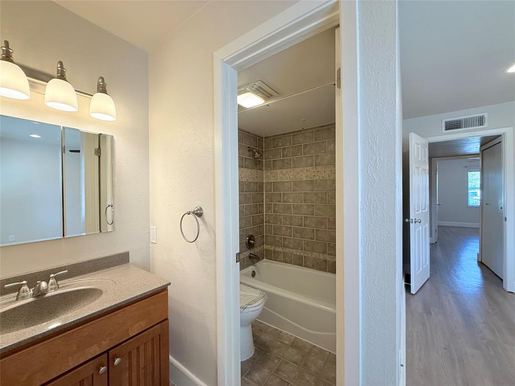 Bathroom featuring a wood-finish vanity with a stone-composite countertop, integrated sink, three-light wall fixture, and a tub-shower enclosure with tiled surround