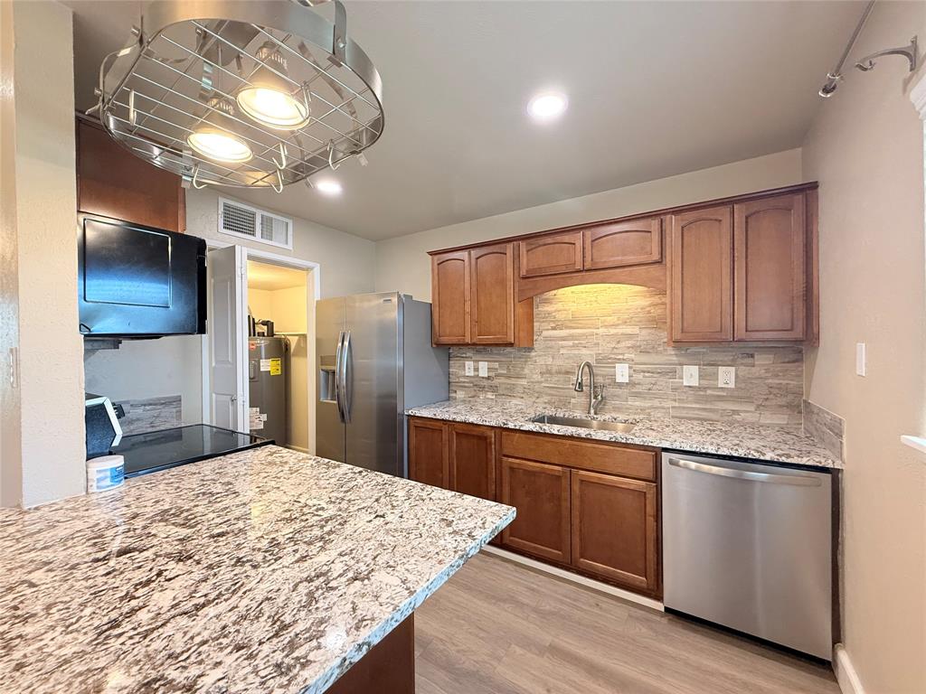 Kitchen featuring stained wood cabinetry, stone-patterned countertops, a stacked stone tile backsplash, stainless steel appliances, and a circular metal pot rack fixture