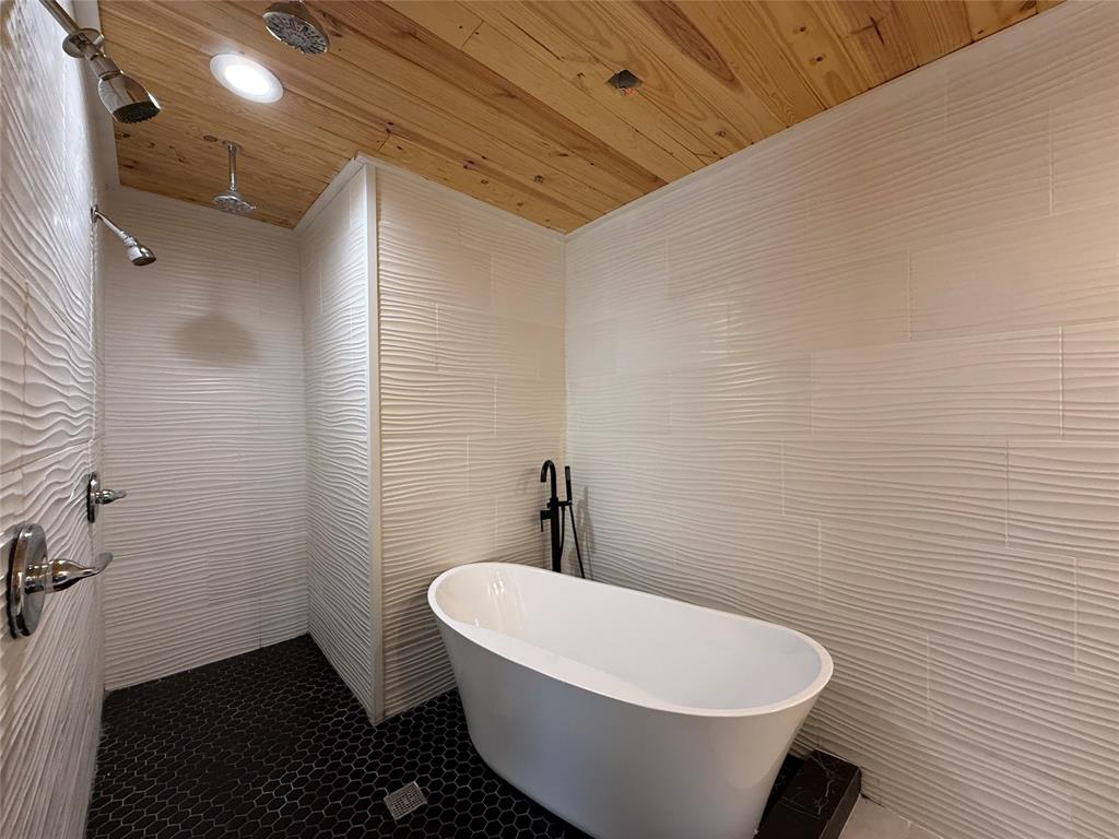 Freestanding soaking tub with matte black fixtures, white textured wall tile, wood-plank ceiling, and black hexagonal floor tile, complemented by dual overhead shower heads