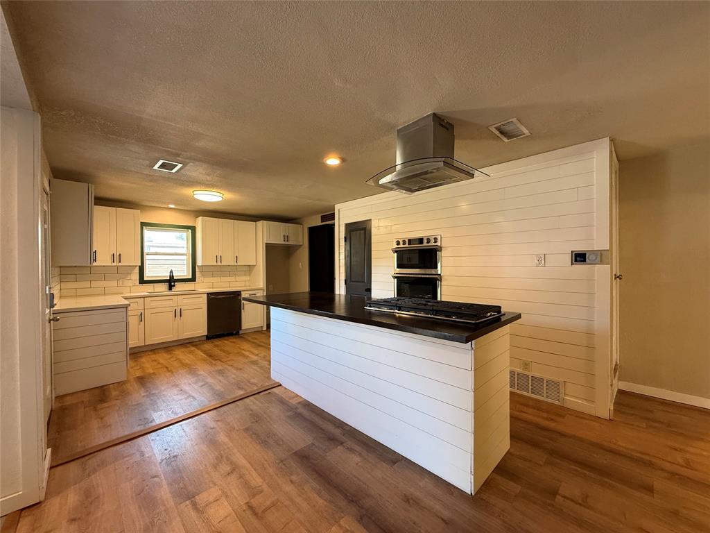 Kitchen featuring white shaker-style cabinetry, wood-finish flooring, a central island with a dark countertop and inset cooktop, a stainless steel range hood, and a built-in double wall oven