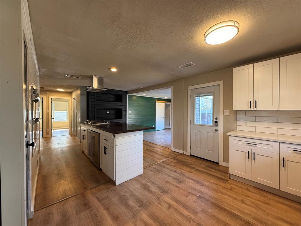 Kitchen featuring white shaker cabinetry, wood-finish flooring, a central island with a built-in cooktop, stainless steel ventilation hood, and white subway tile backsplash