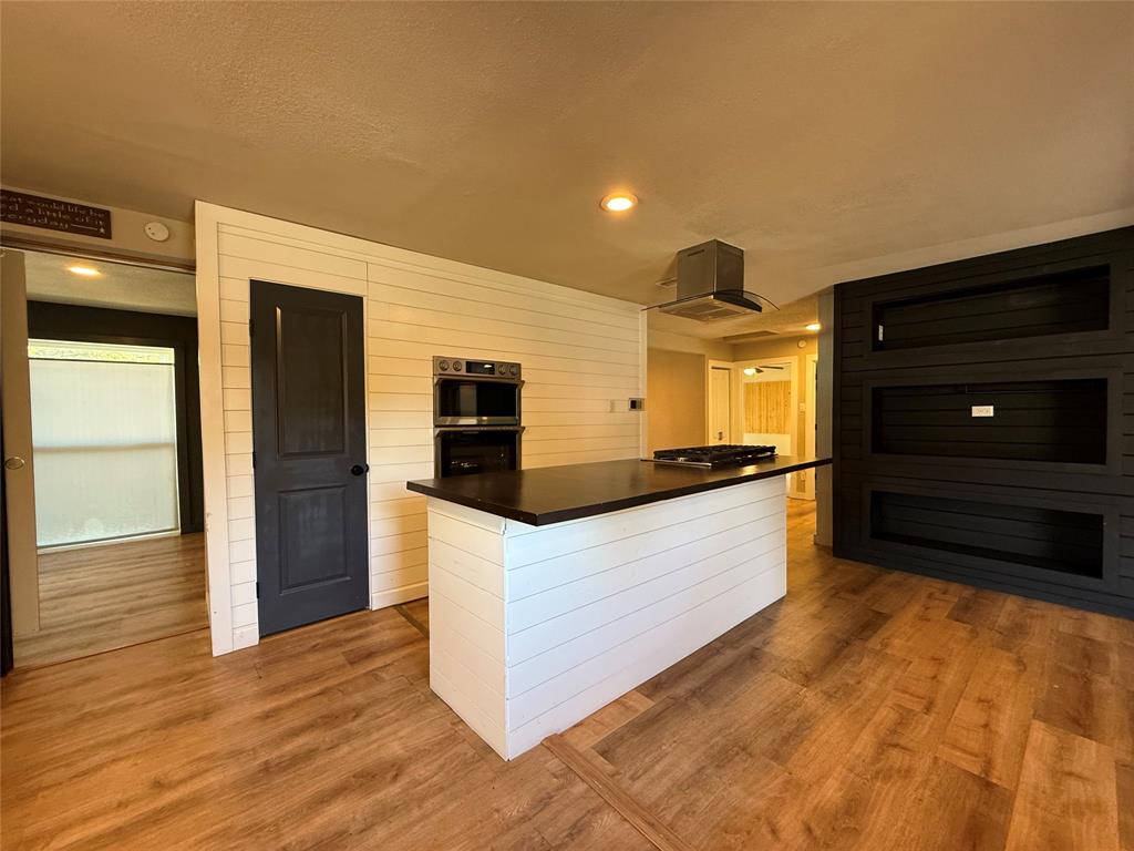 Kitchen featuring a central island with a dark countertop, stainless steel wall-mounted oven, stainless steel range hood, white horizontal shiplap wall paneling, and wood-finish flooring