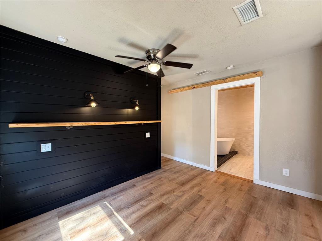 Black shiplap accent wall with built-in wood shelving and dual wall-mounted light fixtures, wood-finish flooring, ceiling fan, and natural wood barn door hardware
