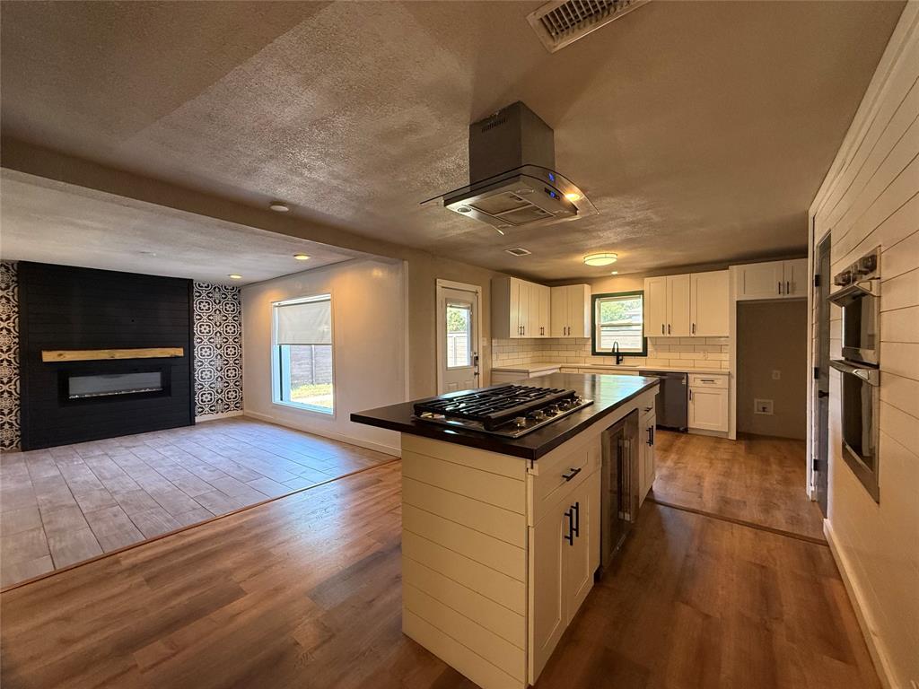 Kitchen featuring a shiplap-wrapped island with an integrated gas cooktop, built-in double wall oven, stainless steel range hood, and white cabinetry paired with a black fireplace accent wall