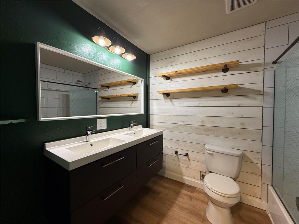 Dual-sink vanity with a rectangular countertop, white plank-style wall detailing, built-in wooden shelving, wood-finish flooring, and a large wall-mounted mirror