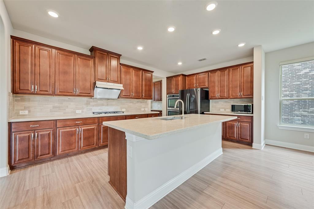 Kitchen featuring wood cabinetry, stone-finish countertops, a central island with an integrated sink, stone-finish tile backsplash, and wood-finish flooring