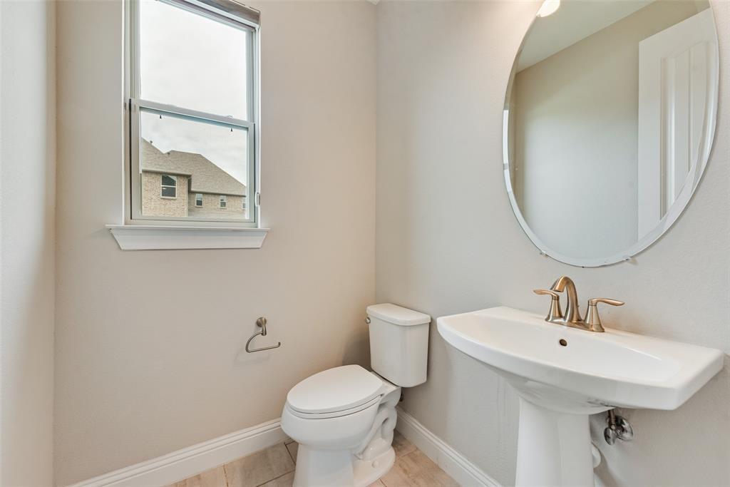 Pedestal sink with metallic faucet, oval wall-mounted mirror, white ceramic toilet, single-hung window, and light-tone tile flooring