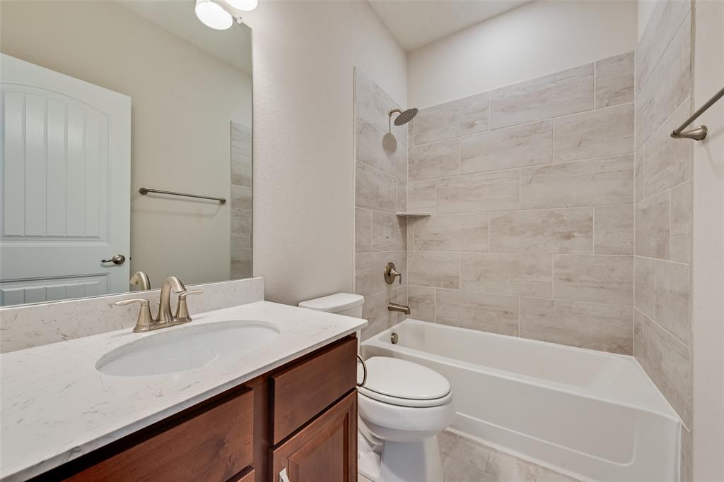 Bathroom featuring a wood-finish vanity with a stone-composite countertop, undermount sink, porcelain tile shower surround, and brushed metal fixtures