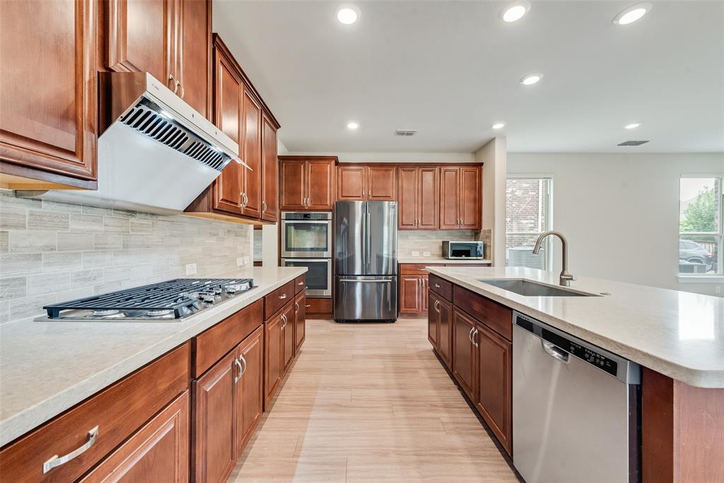Kitchen featuring custom cabinetry, stone-finish countertops, stainless steel appliances including a built-in double oven and gas cooktop, tile backsplash, and a central island with a stainless steel sink
