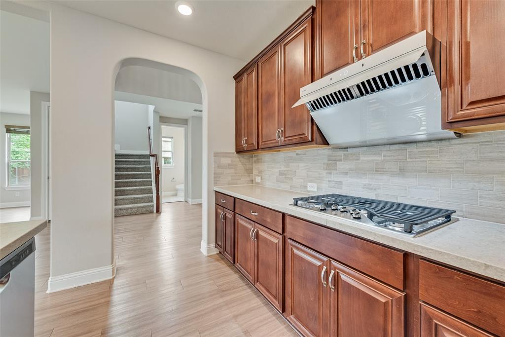 Kitchen featuring wood-finish cabinetry, stone-style countertops, linear tile backsplash, stainless steel gas cooktop, and a matching range hood