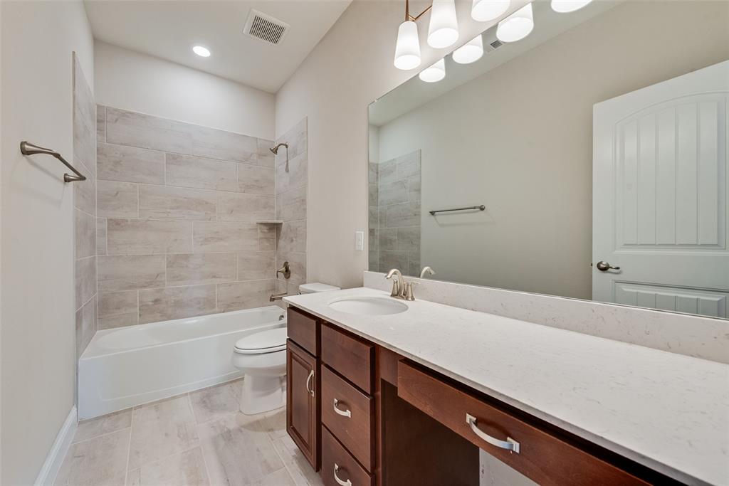 Bathroom featuring a dual-drawer vanity with a stone-finish countertop, undermount sink, porcelain tile tub surround, and oversized wall-to-wall mirror with multi-bulb vanity lighting