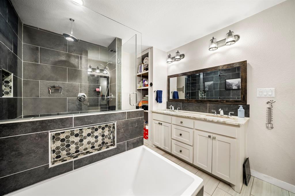 Bathroom featuring a glass-enclosed shower with large-format tile, a built-in recessed niche with hexagonal mosaic accents, a double-sink vanity with a light-toned stone countertop, and built-in floor-to-ceiling shelving