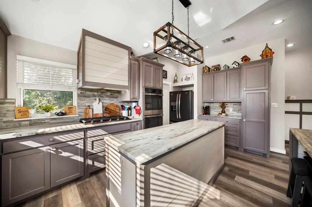 Kitchen featuring a central island with a stone countertop, grey shaker-style cabinetry, horizontal-plank range hood surround, stone-tile backsplash, and rectangular lantern-style pendant lighting