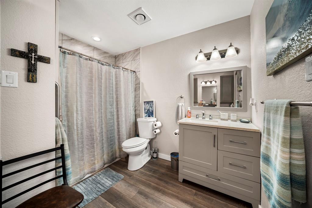 Bathroom featuring a shaker-style vanity with stone-finish countertop, wood-finish flooring, three-light wall-mounted fixture, and tile-surround shower