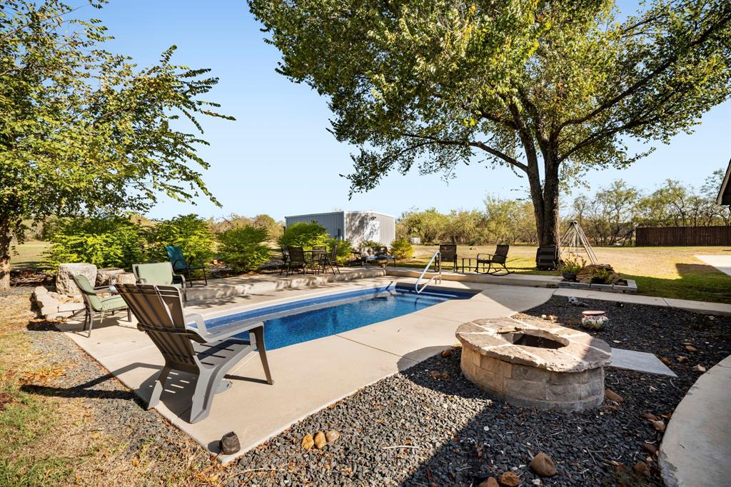 Inground swimming pool with concrete decking, stone-surround fire pit, mature shade trees, and detached storage structure