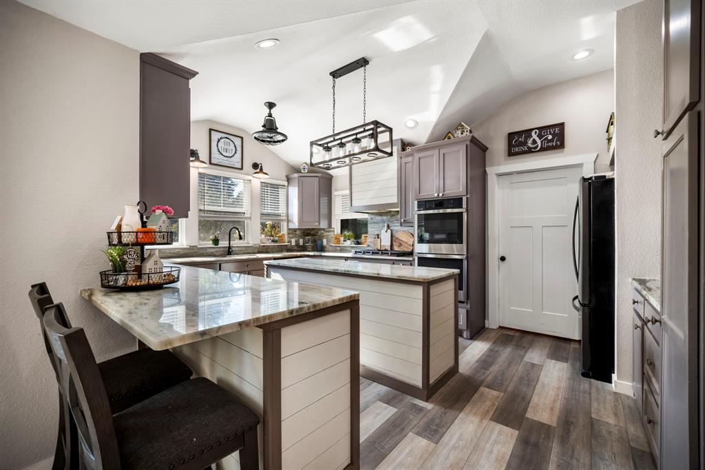 Kitchen featuring shiplap-accented islands, stone-finish countertops, wood-finish flooring, built-in double ovens, and decorative pendant lighting
