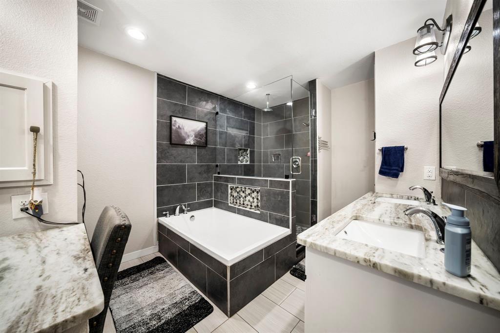 Dual-sink vanity with stone countertop, soaking tub with tile surround, glass-enclosed shower, and recessed lighting