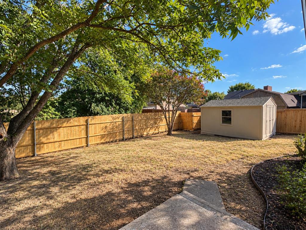 Backyard featuring a perimeter wood fence, detached storage structure, concrete pathway, mature trees, and ground cover