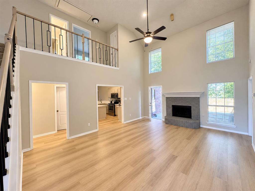 Double-height living area featuring wood-finish flooring, a brick fireplace with a decorative mantel, multiple large windows, and a metal-baluster staircase railing