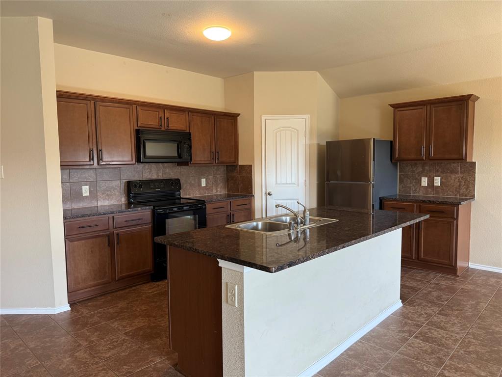Kitchen featuring wood cabinetry, a central island with a dual-basin sink, stone-finish countertops, tile backsplash, and tile flooring
