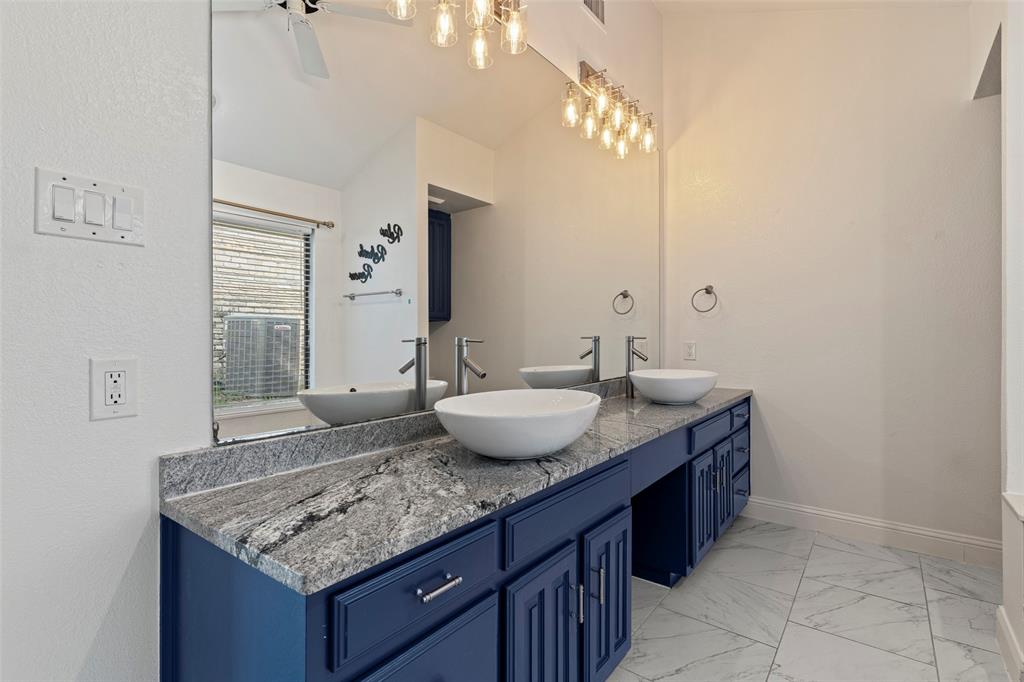 Dual vanity featuring navy blue cabinetry, stone-finish countertops, white vessel sinks, and tiered glass-shade light fixtures, complemented by large-format floor tile