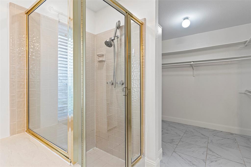 Walk-in shower featuring a metallic frame, hand-held showerhead, and tiled surround, paired with a storage area finished with marble-patterned tile flooring and built-in shelving