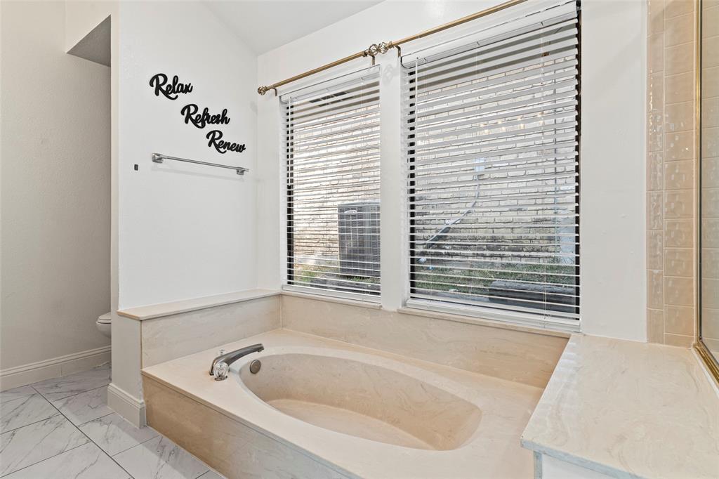 Built-in soaking tub with surround stone-finish deck, dual windows with horizontal blinds, and large-format tile flooring