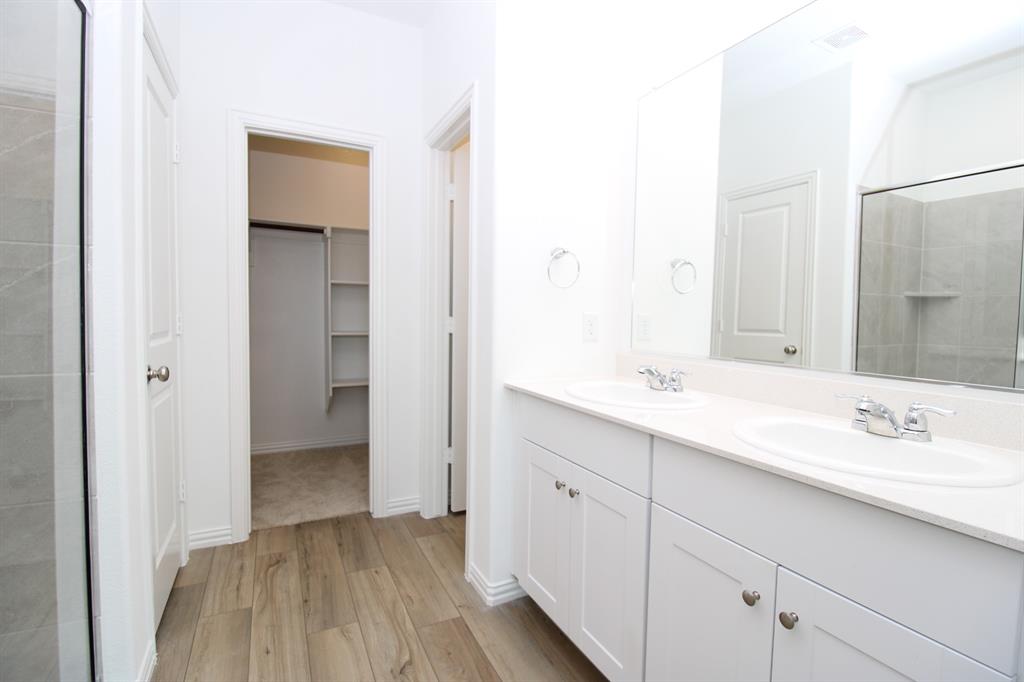 Dual-sink vanity with light-toned countertop, wood-finish flooring, full-width mirror, walk-in closet with built-in shelving, and glass-enclosed shower with tile surround