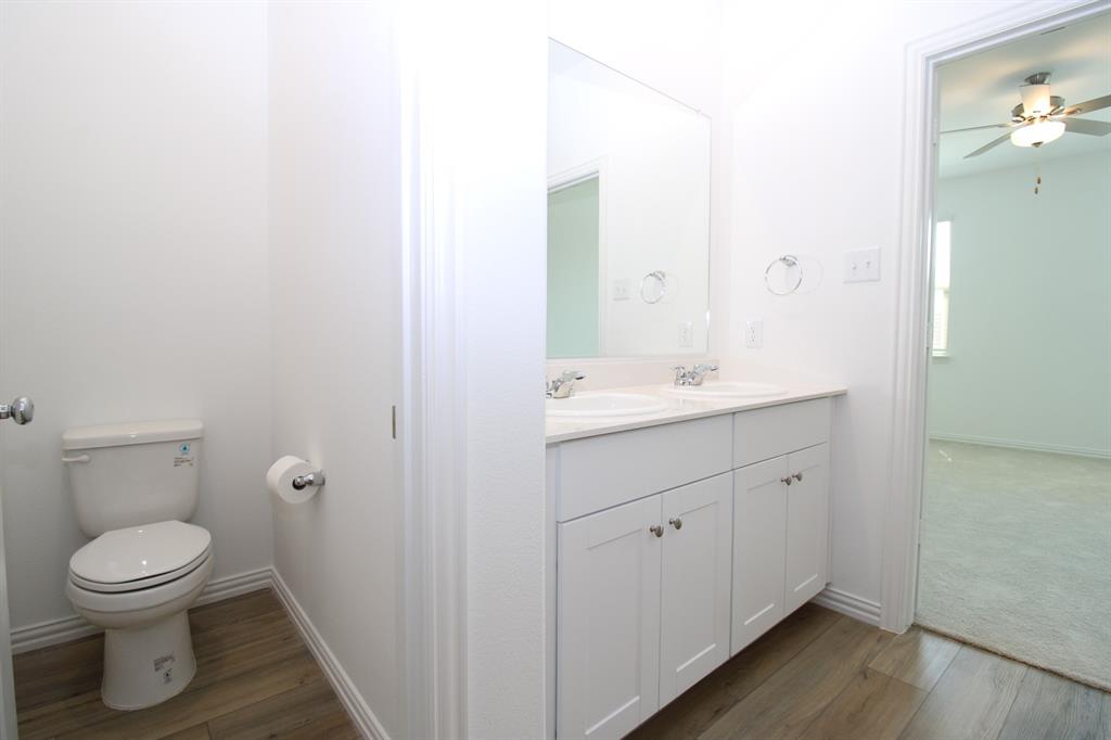 Dual-sink vanity with white shaker-style cabinetry, light-toned countertop, wood-finish flooring, and wall-mounted mirror