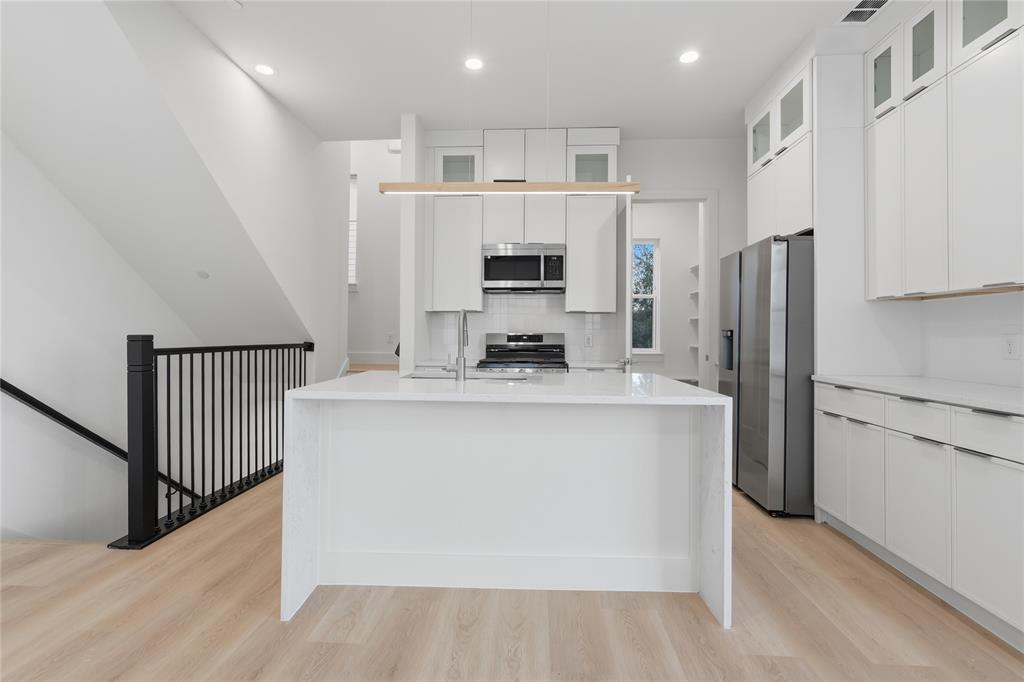 Kitchen featuring an expansive waterfall-edge island, white shaker-style cabinetry with glass-front uppers, stainless steel appliances, and wood-finish flooring