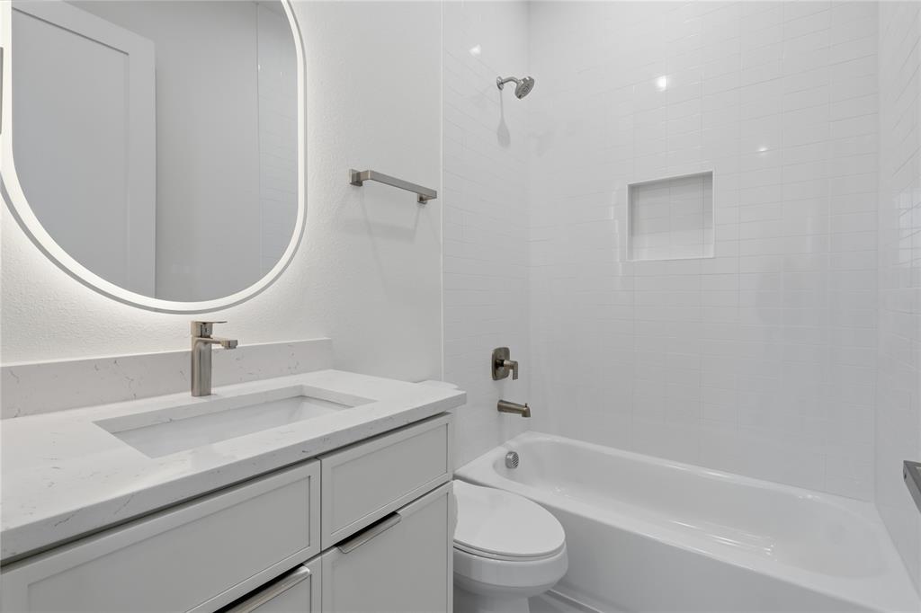 Bathroom featuring a vanity with a quartz countertop, integrated rectangular sink, backlit oval mirror, and a bathtub with a tiled surround and built-in niche